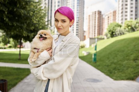 cheerful purple-haired woman holding funny pomeranian spitz in hands and looking at camera on street