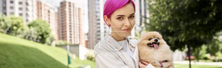 joyful purple-haired woman with cute pomeranian spitz in hands looking at camera on street, banner