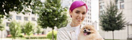 trendy purple-haired woman with pomeranian spitz in hands smiling at camera on urban street, banner