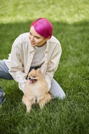 cheerful purple-haired woman cuddling funny pomeranian spitz and looking away on green grass in park
