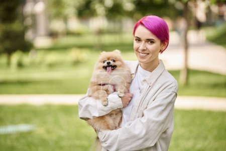 happy and stylish woman holding pomeranian spitz in hands and looking at camera outdoors, enjoyment