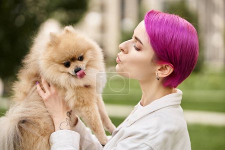 trendy woman with purple hair pouting lips while holding adorable pomeranian spitz outdoors