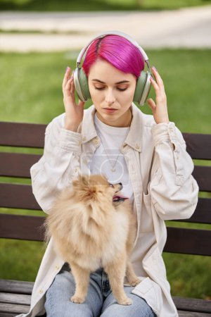 lila haarige Frau hört Musik in drahtlosen Kopfhörern, während sie mit Hund auf Parkbank sitzt