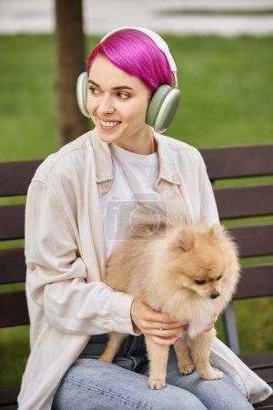 mujer alegre con spitz pomeraniano escuchando música en auriculares y mirando hacia otro lado en el parque