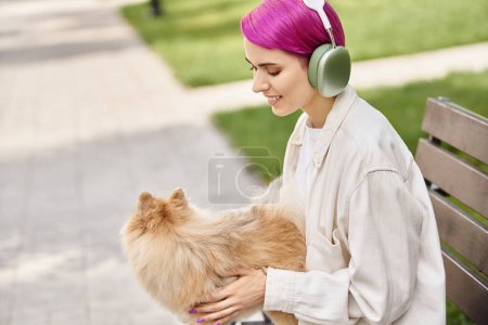 joyful woman listening music in headphones and hugging fluffy pomeranian spitz on bench in park
