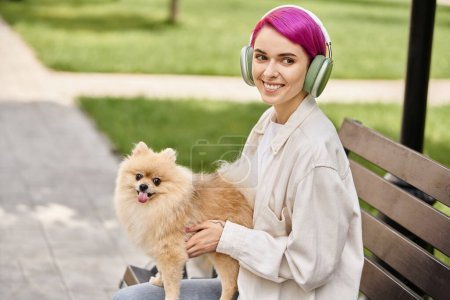carefree purple-haired woman in headphones sitting on bench with furry friend and smiling at camera