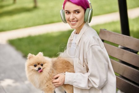 pleased purple-haired woman sitting in park with pomeranian spitz and listening music in headphones