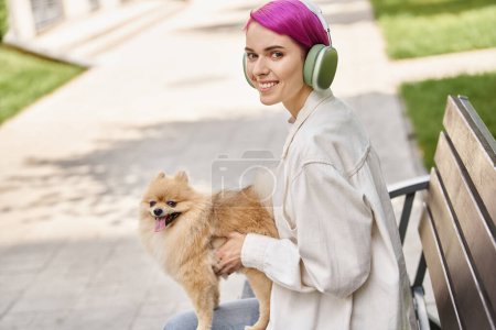 delighted woman with purple hair sitting in park with dog and listening music in headphones