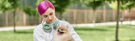 female pet owner with purple hair and headphones holding cute pomeranian spitz in hands, banner