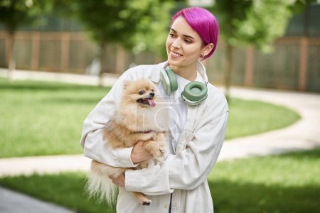 smiley femme aux cheveux violets avec écouteurs marchant avec adorable chien de race dans les mains à l'extérieur