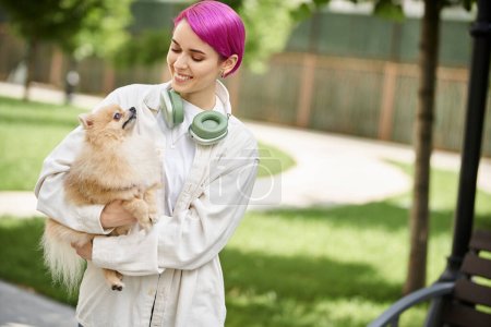 positive purple-haired woman with headphones holding pampered pomeranian spitz in hands outdoors