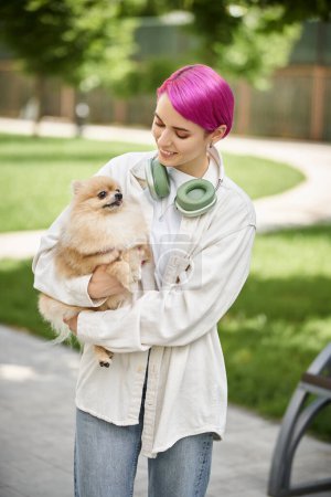 pleased woman with purple hair and headphones standing with loveable pomeranian spitz in hands