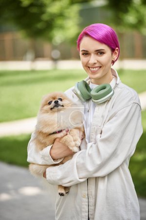 delighted woman with purple hair and headphones holding furry friend and looking at camera outdoors
