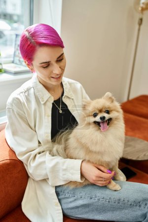 sonriente mujer de pelo morado sentado con spitz pomeranian divertido en moderno hotel de perros, servicio de mascotas