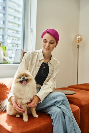 happy woman with purple hair sitting in dog hotel and embracing pomeranian spitz, pet accommodation