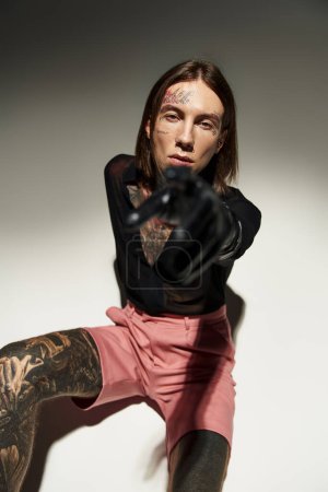 Téléchargez les photos : Jeune homme avec des tatouages assis sur le sol pointant la main dans un gant en cuir à la caméra, concept de mode - en image libre de droit