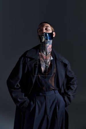 gut aussehender Mann mit Tätowierungen in geschnürter stylischer Maske posiert auf schwarzem Hintergrund, Modekonzept