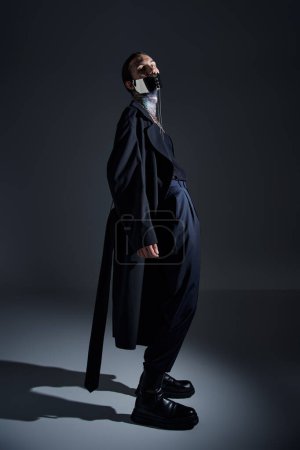Téléchargez les photos : Bel homme élégant en manteau long noir et masque laqué futuriste posant et regardant la caméra - en image libre de droit