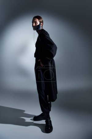 vertical shot of stylish young man in black attire with laced mask surrounded by lights, fashion