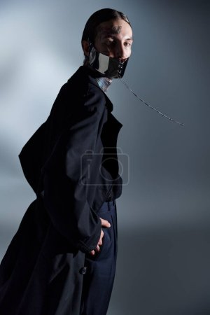 gutaussehender Mann mit Tätowierungen in futuristischer Maske und schwarzer Kleidung, der in die Kamera schaut, Modekonzept