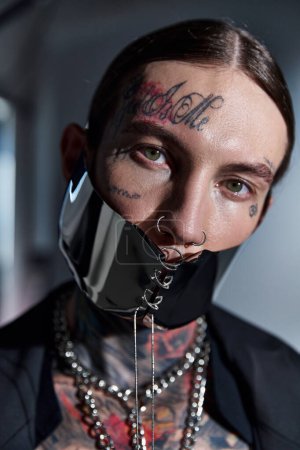 Porträt eines jungen, stylischen Mannes mit Tätowierungen in futuristischer Maske, der in die Kamera blickt, Modekonzept