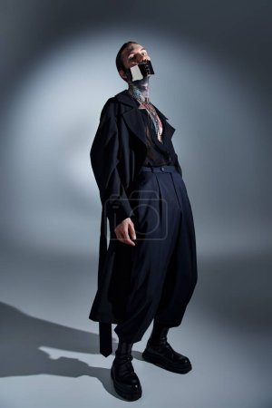 hübsches sexy männliches Modell mit Tätowierungen und futuristischer Maske im schwarzen Mantel, der seinen Körper leicht beugt