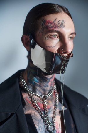 Porträt eines sexy jungen Mannes mit Tätowierungen im Gesicht mit Schnürmaske und Accessoires, die wegschauen