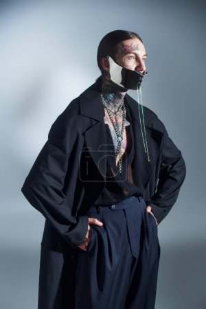 handsome stylish man with futuristic laced mask posing with hands in pockets looking away, fashion