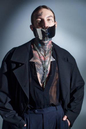 vertical shot of alluring young man with tattoos and laced mask with accessories looking at camera