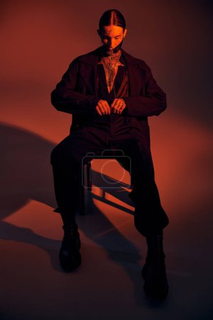 sexy junger Mann in futuristischer Schnürmaske mit stylischen Accessoires auf Stuhl sitzend, Modekonzept