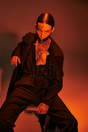 handsome alluring man in futuristic attire sitting on chair and looking at camera, fashion concept