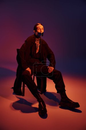 stylish young man in black coat and laced stylish mask sitting on chair looking at camera, fashion