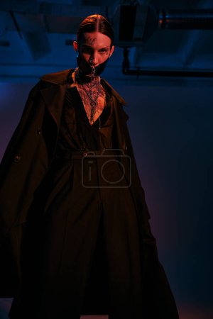 vertikale Aufnahme eines jungen, stylischen Mannes in schwarzer futuristischer Kleidung, der im dunklen Licht posiert, Modekonzept
