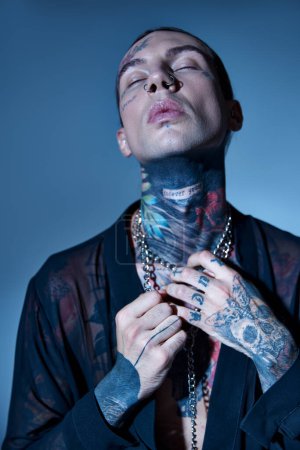 hombre atractivo atractivo con tatuajes elegantes posando con los ojos cerrados tocando sus accesorios, moda