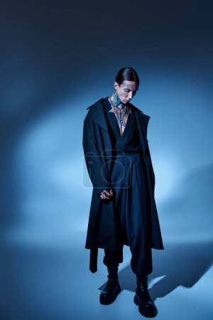 vertical shot of handsome young man with tattoos posing in black voguish coat and looking down