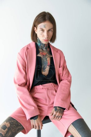handsome man in stylish outfit with tattoos sitting on chair and looking at camera, fashion concept