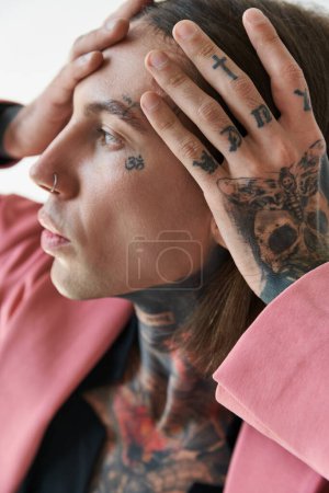 Téléchargez les photos : Portrait de jeune homme à la mode avec des tatouages sur le visage et perçage avec les mains sur le visage détournant les yeux - en image libre de droit