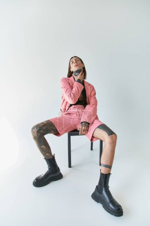 Foto de Hombre sexy con estilo en chaqueta rosa y pantalones cortos sentados en la silla y mirando a la cámara con la mano en el cuello - Imagen libre de derechos