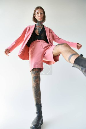 Téléchargez les photos : Sexy jeune modèle masculin avec des tatouages dans une tenue vibrante élégante avec jambe levée regardant la caméra - en image libre de droit