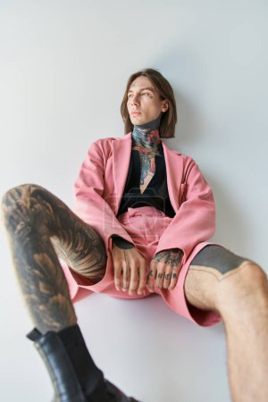 Téléchargez les photos : Beau jeune homme avec des tatouages et piercing en veste rose détournant les yeux, concept de mode - en image libre de droit