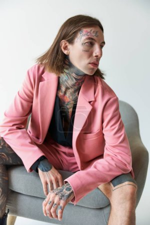 Téléchargez les photos : Plan vertical de jeune homme séduisant avec des tatouages relaxant sur la chaise et détournant les yeux, concept de mode - en image libre de droit