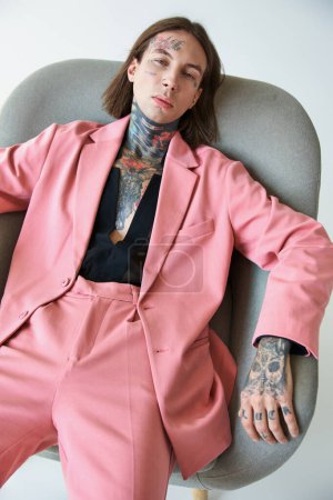 Téléchargez les photos : Bel homme sexy en tenue voguish avec des tatouages en regardant la caméra tout en se relaxant sur la chaise, la mode - en image libre de droit