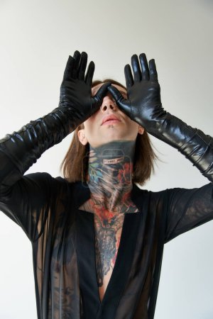 Téléchargez les photos : Jeune homme séduisant avec tatouages et perçage avec les mains en gants de cuir sur le visage, concept de mode - en image libre de droit
