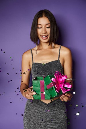 excitada mujer asiática en partido vestido celebración envuelto regalo de Navidad cerca de caer confeti en púrpura