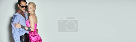Téléchargez les photos : Femme blonde en robe rose câlin avec petit ami tatoué dans des lunettes de soleil sur gris, bannière Nouvel An - en image libre de droit