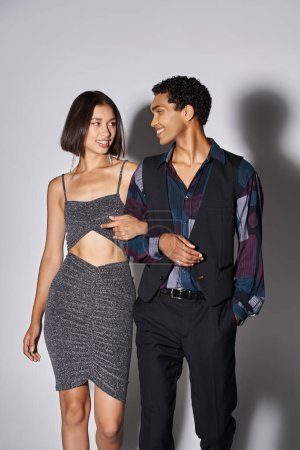 glückliche asiatische Frau im Partykleid mit afrikanisch-amerikanischem Mann auf grau, Neujahr 2024
