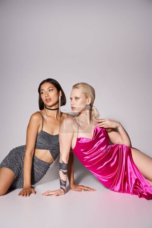two interracial female friends in dresses sitting on grey backdrop, New Year party concept