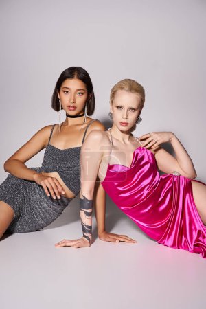 two pretty multiethnic women in dresses sitting on grey backdrop, Merry Christmas party concept