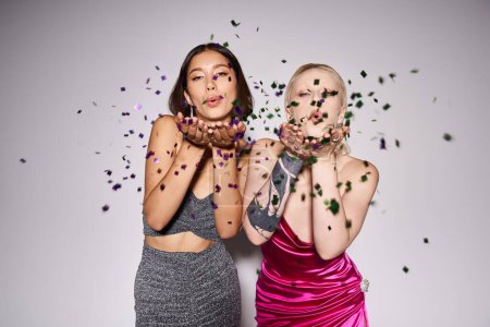 two multiethnic women in party dresses blowing confetti on grey background, New Year party concept