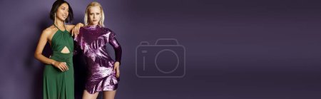 happy asian woman posing with blonde friend, standing together in party dresses on purple, banner
